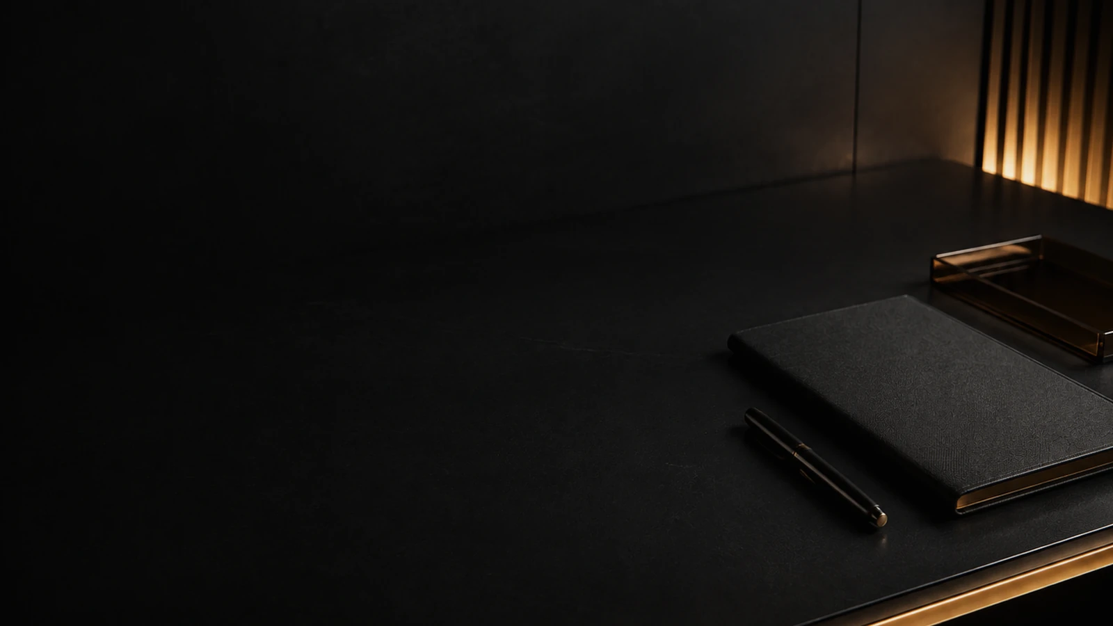 An almost empty dark desk holding one closed black notebook and a capped pen, lit only by a warm slatted panel at the far right.