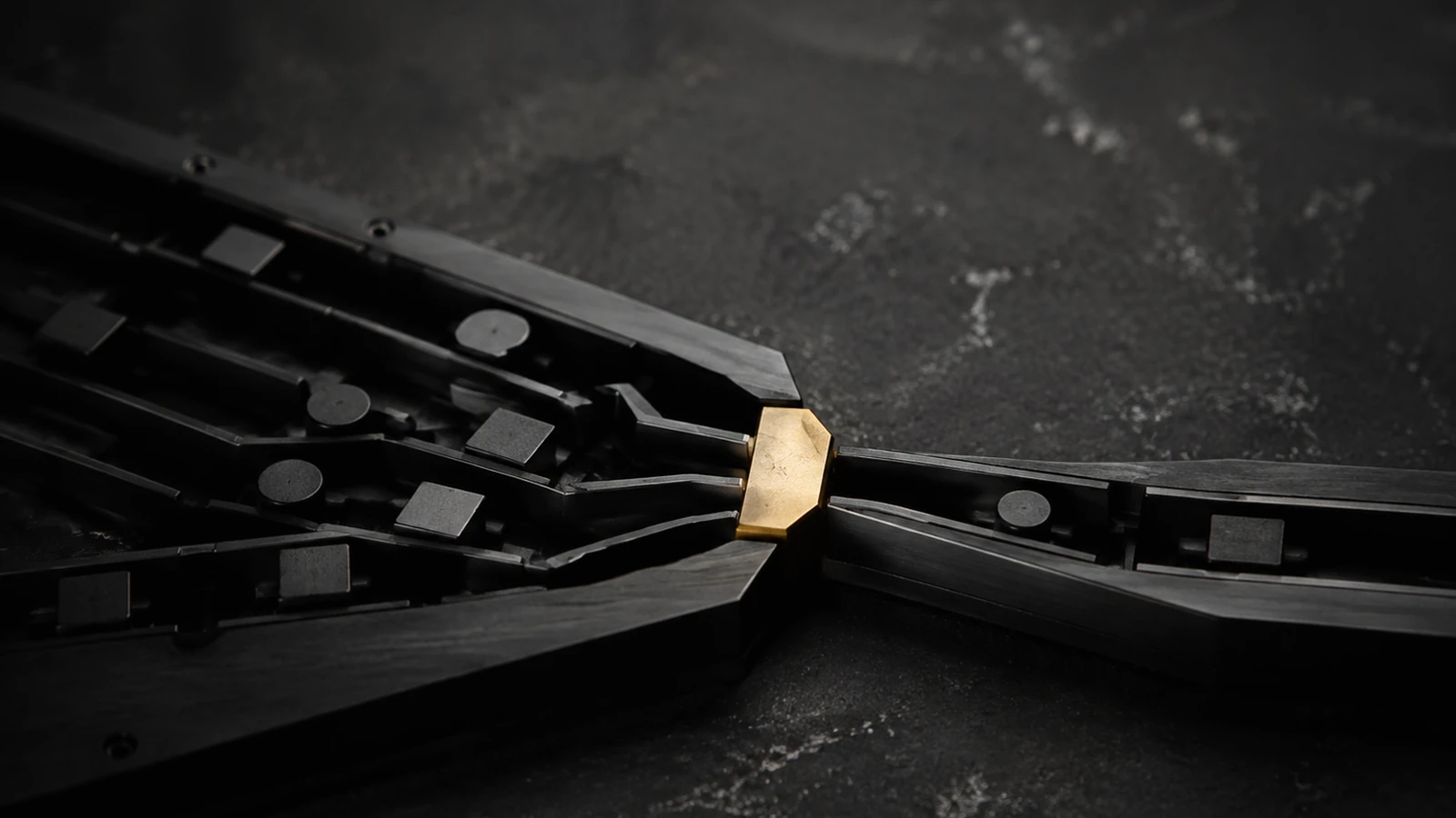 Close view of several matte black machined arms converging from the left onto one brass wedge, with a single arm carrying on past it to the right.